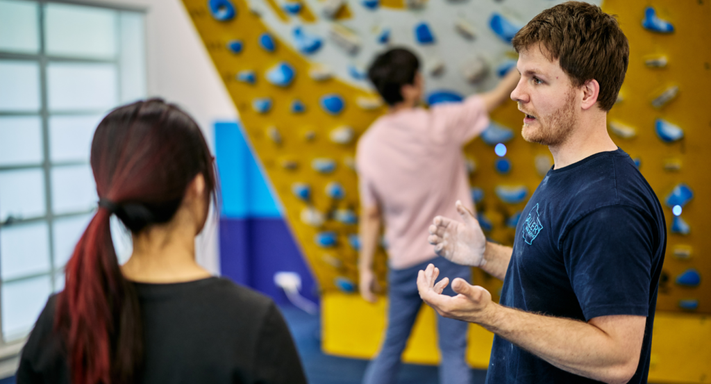 Coach Max speaks to a woman in front of the Moonboard in a climbing gym.